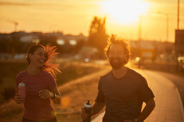 Two joggers share laughter as they run along a paved trail during sunset, embodying a healthy lifestyle and the joy of exercising together outdoors.