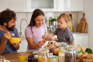 A happy family shares breakfast at home, with a parents smiling and a young child playfully feeding a stuffed animal. The kitchen is bright and welcoming, filled with warmth.