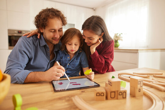 A family of three is gathered around a tablet on a wooden table in their bright kitchen. They are engaged in a fun drawing activity, smiling and sharing moments of joy.