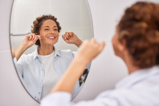 A smiling woman performs her dental care by flossing her teeth in a well-lit bathroom. She enjoys her morning routine while looking in the mirror, showing good hygiene habits. - Powered by Adobe