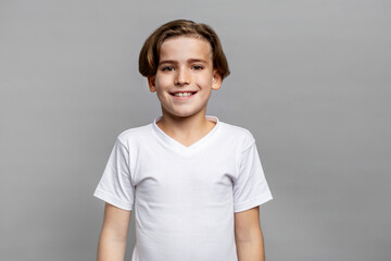 Smiling brunette boy with brown eyes posing for studio portrait, looking at camera in white t-shirt, isolated on neutral gray background, expressing happiness and positive emotion