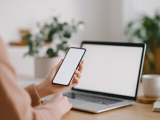 Woman holding a smartphone with a blank white screen, sitting at a desk and working remotely, receiving information, or multitasking with a laptop also featuring a blank display