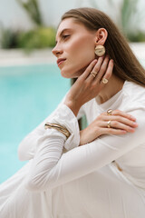 Profile close-up of a woman in a white outfit touching her face, with a soft turquoise pool in the background