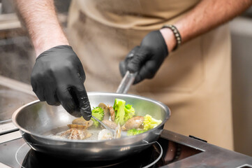 A chef wearing black gloves stirs clams and broccoli in a stainless steel pan on a stovetop, with steam rising as they cook. Focus on hands and food preparation