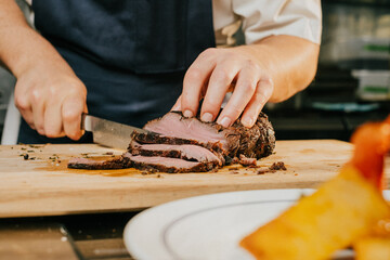 A chef is cutting a piece of meat on a wooden cutting board. The knife is positioned above the meat, and the chef's hand is holding the knife