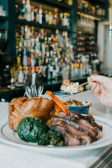 A person is eating a plate of food that includes a steak, potatoes, and vegetables. The plate is on a table in front of a bar