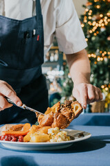 A man is serving food on a plate. The plate has a variety of food items, including meat, vegetables, and potatoes. The man is using a fork to serve the food onto the plate