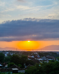 Fototapeta premium Golden sunrise emerges above distant mountains and a calm strait, casting warm light over Banyuwangi’s eastern horizon as the town slowly brightens beneath layered clouds.