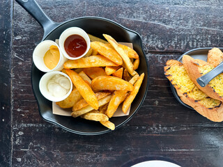 Golden brown seasoned potato wedges served in a black skillet with ketchup, mayonnaise, and mustard sauces, alongside toasted bread with yellow spread.