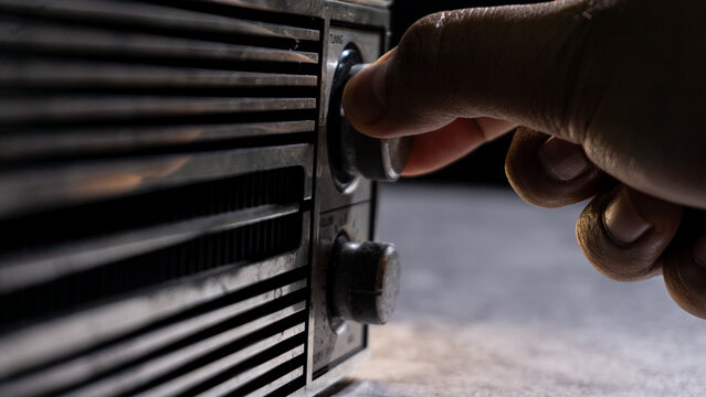 A hand adjusting the tuning knob of a weathered radio, with dramatic shadows highlighting texture and vintage craftsmanship. Suitable for themes of retro sound and manual controls.