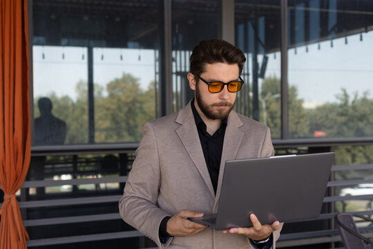 A man is standing on a balcony while using a dark-colored laptop computer