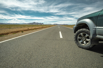 An off-road vehicle with all-terrain tires parked on an asphalt road in the Mongolian steppe. Close-up on the front left wheel against a cloudy blue sky.

