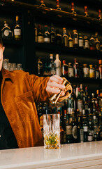 A bartender is pouring a drink into a glass. The bar is full of bottles