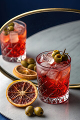 High-quality still life photograph of a cocktail glass filled with red liquid and ice cubes, garnished with green olives and a slice of dried citrus fruit.