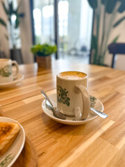 Close-up of a floral coffee cup with frothy milk on a saucer and spoon, set on a wooden café table with toast and natural light in the background.