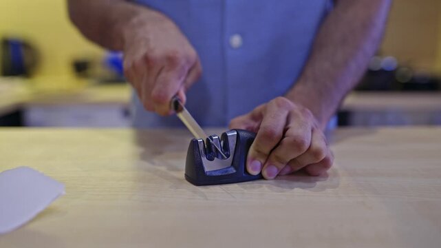 A close-up view shows a man's hands sharpening a kitchen knife with a handhold sharpener on a wooden countertop, accompanied by the distinct grinding and scraping sounds of the process.
