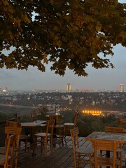 A quiet Belgrade terrace with wooden seating opens to a panoramic dusk view, where warm city lights and distant buildings form a serene contrast against the soft, evening sky.