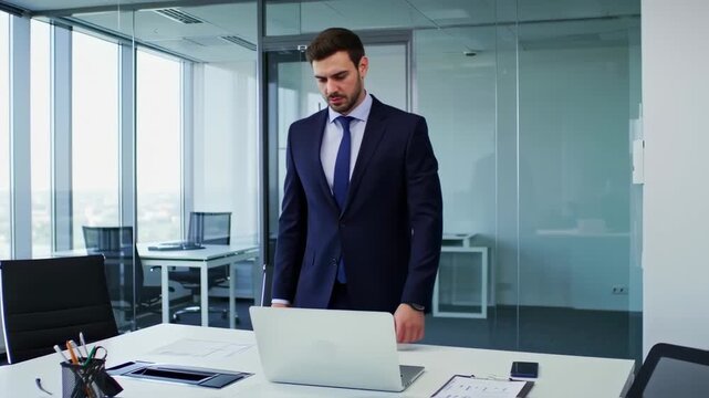 A formally dressed man in an office looks intently at a laptop on a white desk