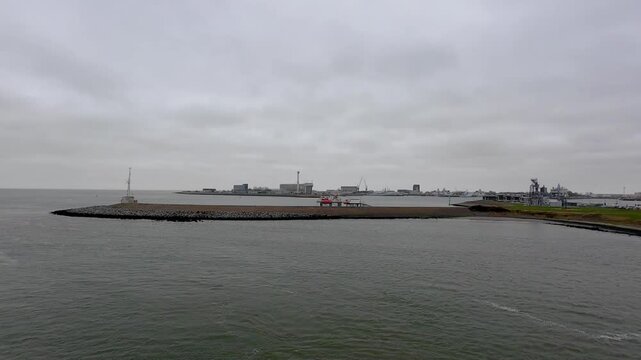 Den Helder, The Netherlands, October 25th, 2024, overcast harbor skyline with breakwater, industrial port buildings and cranes on distant shore, calm green water lapping jetty,
