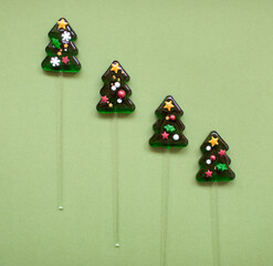 Four green christmas tree shaped lollipops with holiday sprinkles arranged diagonally on a muted green background with copy space, top view