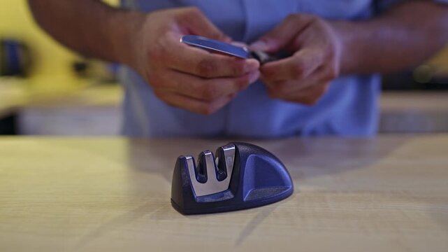 Watch a detailed close-up shot of hands using a manual sharpener to hone a kitchen knife's blade on a wooden surface. This video includes the authentic sounds of the sharpening process.
