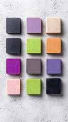 Fototapeta premium A collection of twelve square, handmade soaps arranged in a grid on a textured gray surface. The soaps vary in color, including black, purple, lavender, beige,
