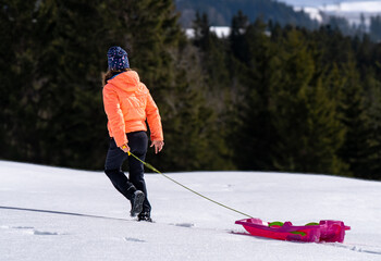 A child in a bright orange jacket and winter hat walks through deep, fresh snow, pulling a pink sled. Forested mountains create a serene winter wonderland background.