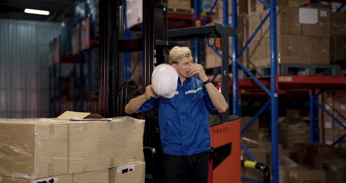 caucasian male warehouse staff in blue uniform holding white safety helmet standing beside cardboard boxes leaning on forklift pallet truck during working break inside logistic storage area