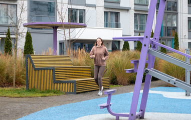 A young woman runs near an outdoor workout area in a residential courtyard. She jogs past fitness