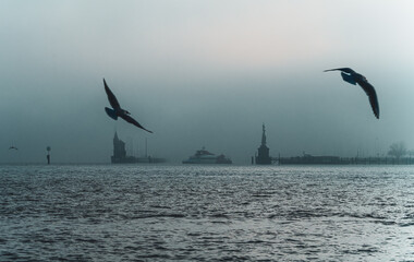 Naklejka premium On a foggy winter day, the ferry departs from the port of Konstanz with the Imperia statue and the lighthouse, while seagulls fly in the wind. Konstanz, Baden-Württemberg, Germany.