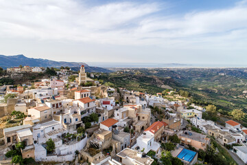 Hilltop village with white houses, red roofs, tall clock tower overlooking vast valley and distant sea, creating bright Mediterranean panorama with layered hills and sunlit rural scenery.