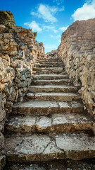 Old stone stairway rising between weathered rock walls under bright sky, creating ancient historic atmosphere with textured stones, warm light, rugged surfaces and inviting path leading upward.
