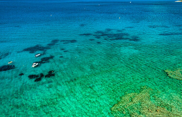 Aerial view of turquoise transparent sea with scattered boats floating above underwater rocks. Calm clear water creating vibrant summer coastal scene with deep and shallow tones.