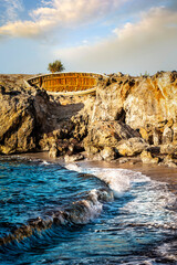 Abandoned wooden boat resting on rocky shore above waves hitting coastline. Warm sunlight highlighting rugged stones, blue sea, golden sky, peaceful Mediterranean seaside landscape.