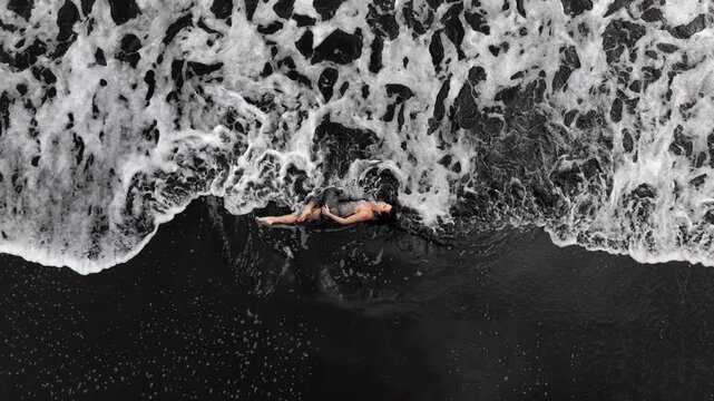 A woman gracefully lies on the ocean shore, shot from above, showing the ebb and flow of the water, the smooth movement of the panorama enhancing the atmosphere of tranquility.