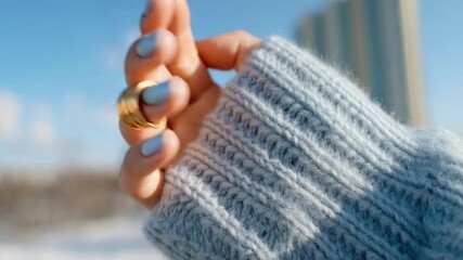 Hand with light blue nails wearing chunky gold rings on a blue knit sweater, outdoors on a sunny day.