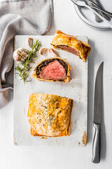 Homemade beef Wellington wrapped in golden puff pastry, sliced to reveal medium-rare beef tenderloin with mushroom duxelles. Styled on a rustic white background with garlic, herbs, and kitchen utensil