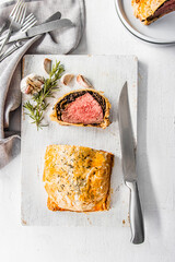 Homemade beef Wellington wrapped in golden puff pastry, sliced to reveal medium-rare beef tenderloin with mushroom duxelles. Styled on a rustic white background with garlic, herbs, and kitchen utensil