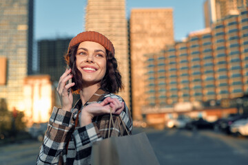 Fototapeta premium Woman shopping on city street, phone to ear and smile, beanie and bag in hand in warm sunlight; authenticity and candid lifestyle portrait with golden glow and emotional storytelling.