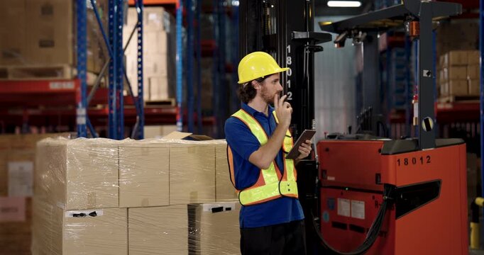 male caucasian warehouse inspector in safety vest and hardhat holding digital tablet checking stock inventory beside stacked cardboard boxes inside logistic cargo distribution facility warehouse - Powered by Adobe