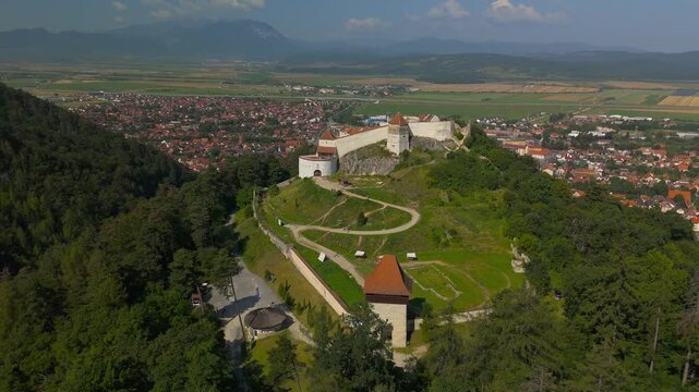Rotating aerial view of the medieval Rasnov fortress in Transylvania, Romania, with the hilltop walls and towers silhouetted against distant Carpathian Mountains and Rasnov town