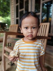A 2-year-old boy is happily eating a donut in front of his house. His expression is cheerful and innocent, with donut crumbs stuck to his mouth. He sits relaxed on a small chair.