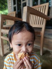 A 2-year-old boy is greedily eating a donut in front of his house. His facial expression is cheerful and innocent, with donut crumbs sticking around his mouth. He sits relaxed on a small chair.