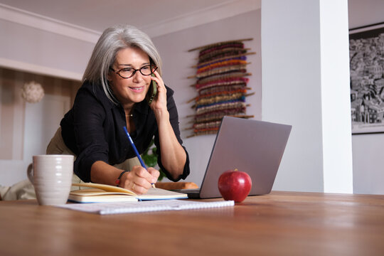 Smiling mature businesswoman working from home office, taking notes while talking on mobile phone and using laptop computer - Powered by Adobe