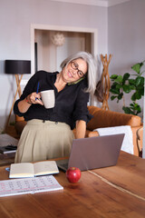Smiling mature businesswoman working from home, drinking coffee and talking on the phone