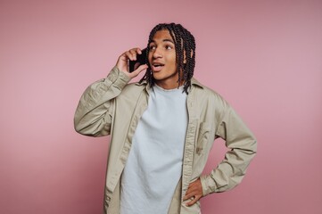 Young man talking on smartphone against a pink background while standing casually with one hand on his hip