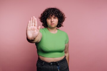 Portrait of a person with curly hair in a green shirt expressing a stop gesture against a pink background
