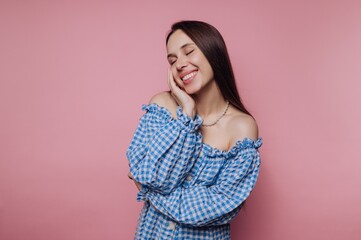 Young woman smiles happily against a pink backdrop while wearing a blue checkered off-shoulder top during a cheerful indoor moment