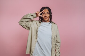 Young man poses playfully against a pink background with a peace sign gesture while wearing a casual outfit