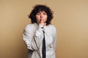 Person making a shushing gesture in a light brown studio setting with a relaxed pose and curly hair